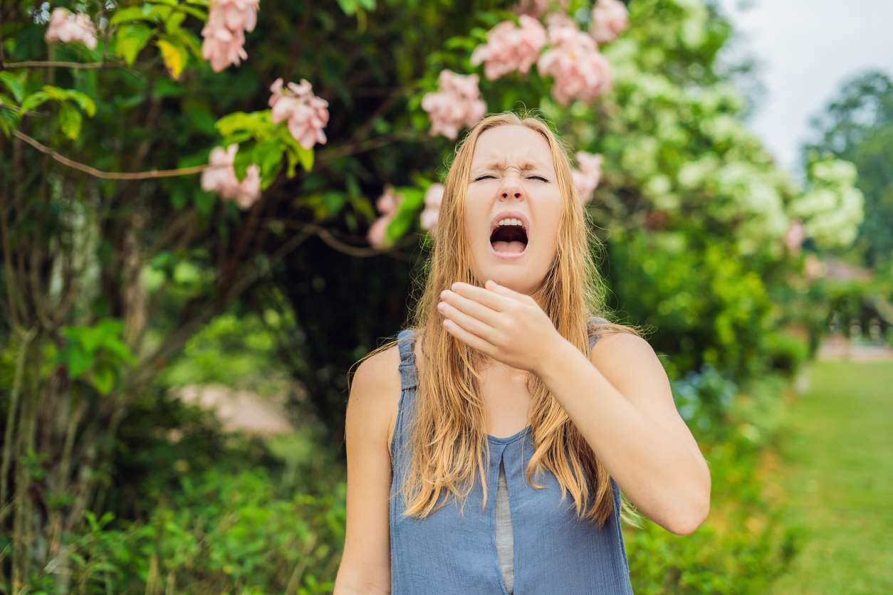 Young woman sneezes in the park against the background of a flowering tree. Allergy to pollen concept.