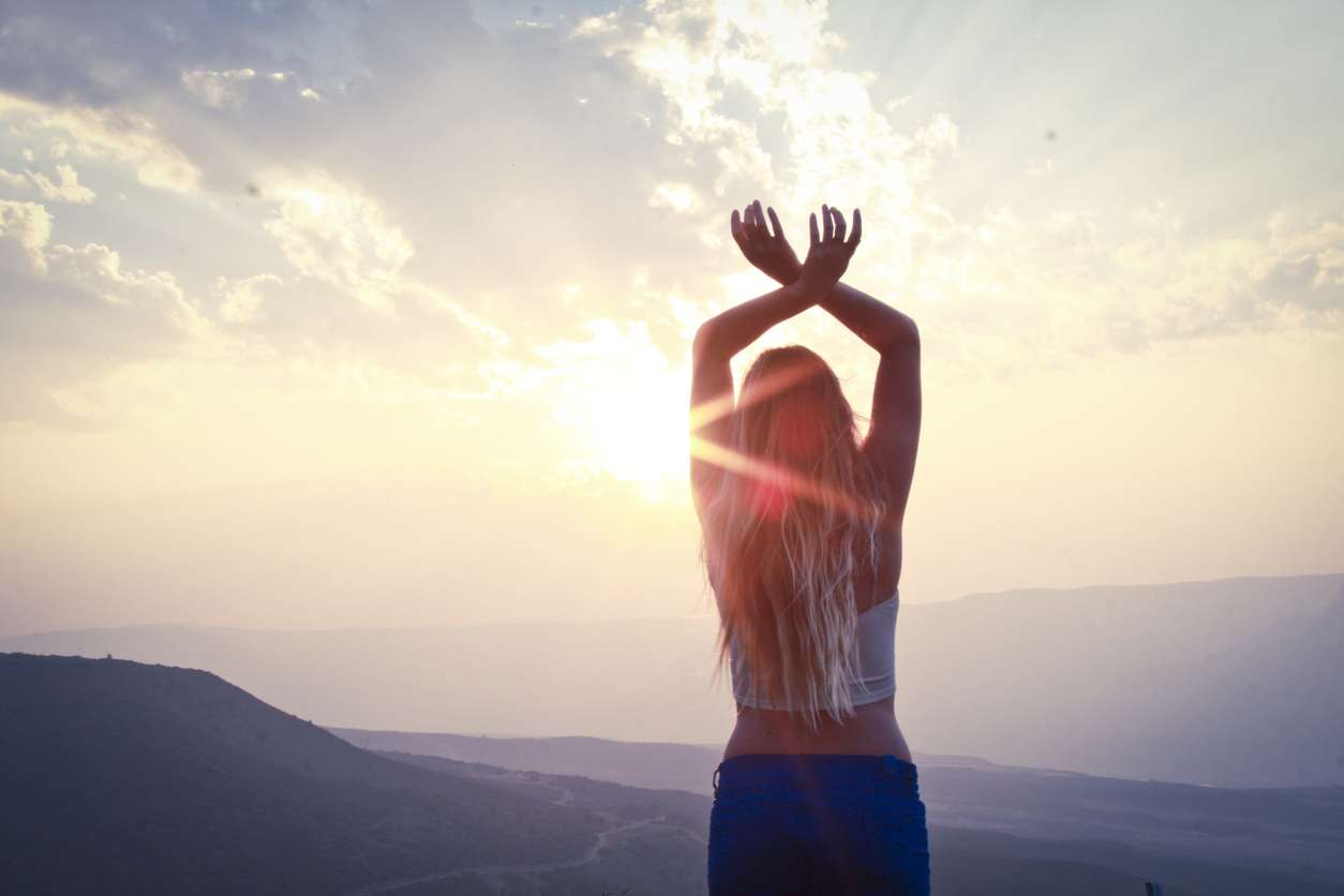 a long blonde haired female with a white cropped top and blue shorts has her hands outstretched above head looking into the sunset.