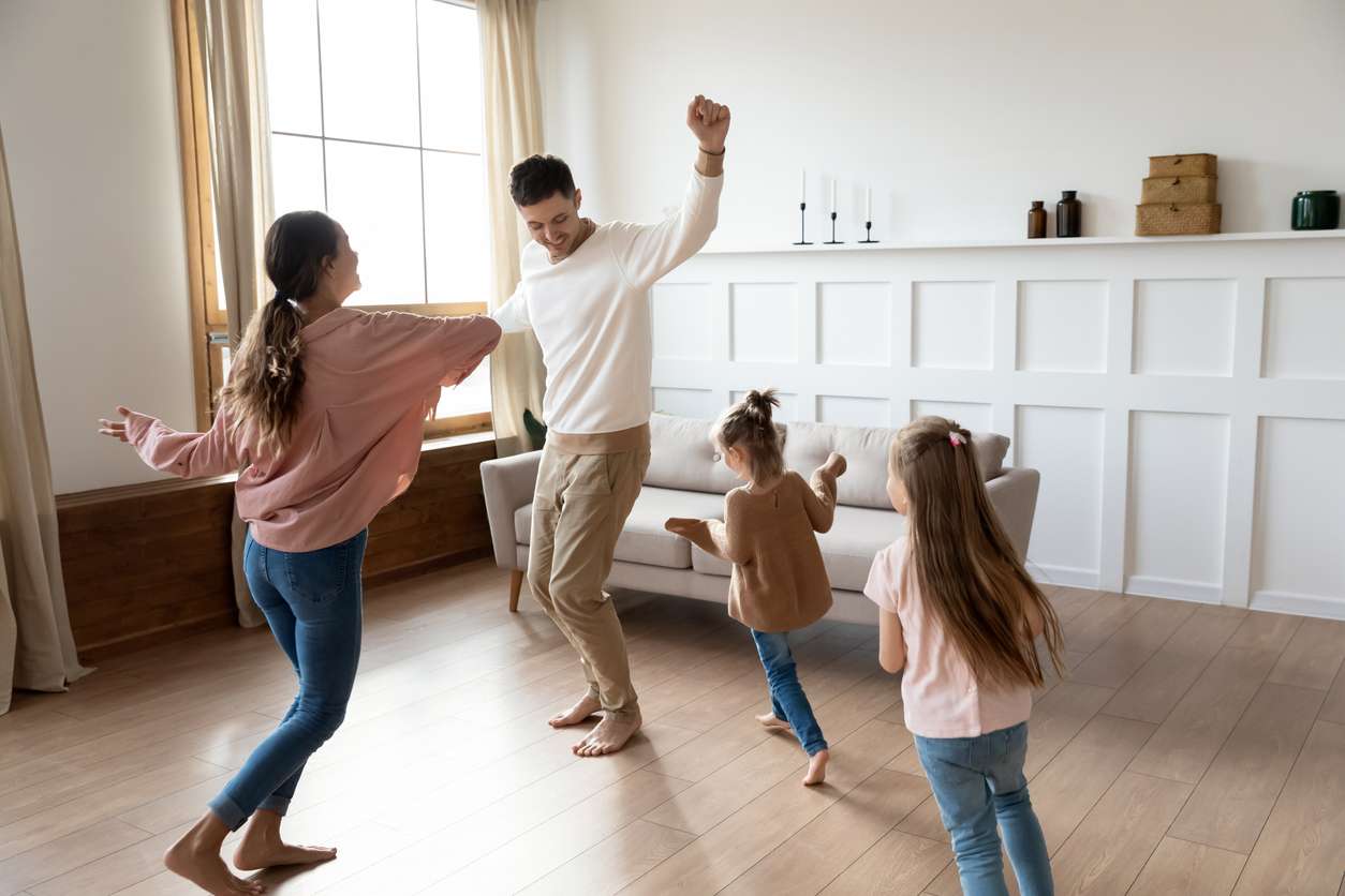 Funny active family of four young adult parents and cute small children daughters dancing together in living room interior, carefree little kids with mum dad having fun laughing enjoy leisure at home